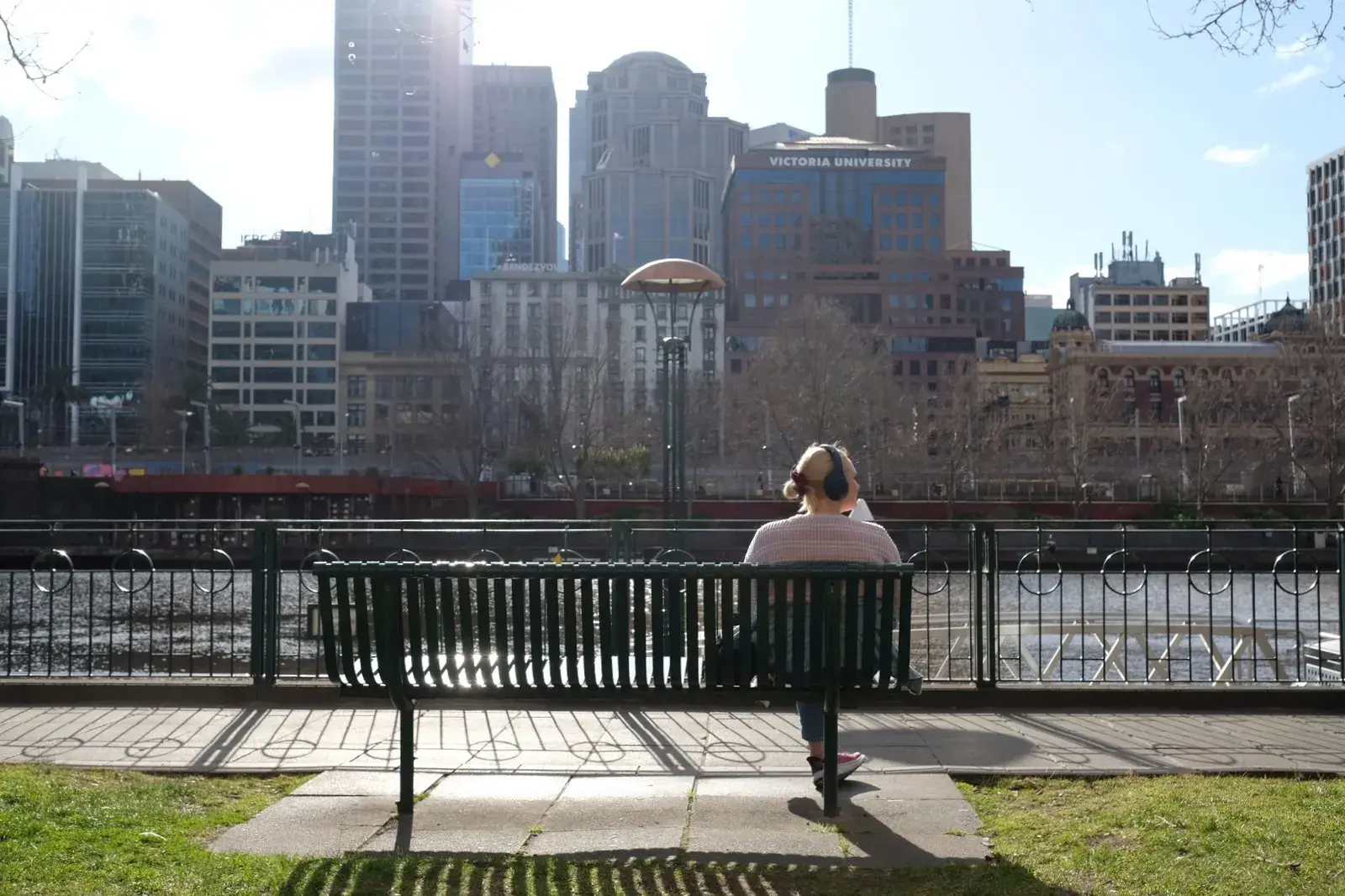 ผู้หญิงใส่หูฟังนั่งบนม้านั่งริมแม่น้ำ Yarra มองตึกสูงใจกลางเมืองเมลเบิร์น
